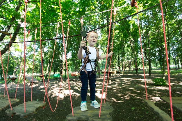 Un niño pequeño con equipo de escalada está caminando por un camino de cuerdas en un parque de aventuras, agarrado a una cuerda y un mosquetón.