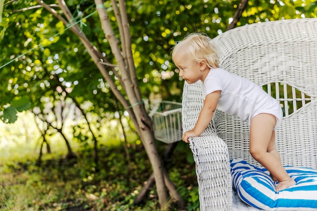 El niño pequeño está jugando en el patio trasero. El bebé disfruta de la infancia, el niño de pelo rubio con un mono blanco para niños se apoya en el costado de una gran silla de lino beige. Disfrutando del sol, momentos familiares