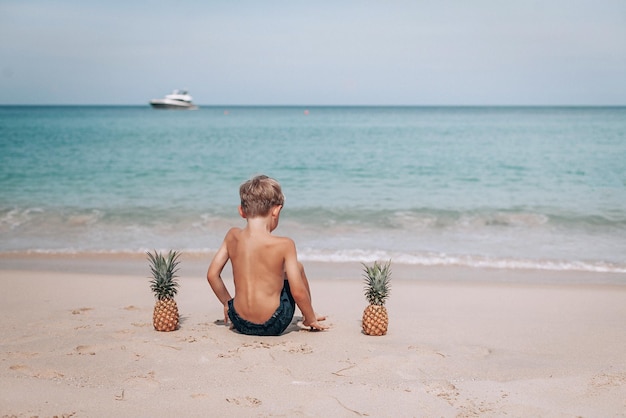 Un niño pequeño está sentado en la orilla del mar y dos piñas yacen cerca. El niño mira al mar