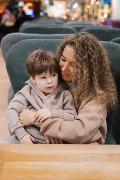 Un niño pequeño está sentado en el regazo de su madre en un centro comercial y de entretenimiento en un café del patio de comidas, una madre se comunica con su hijo pasando tiempo juntos