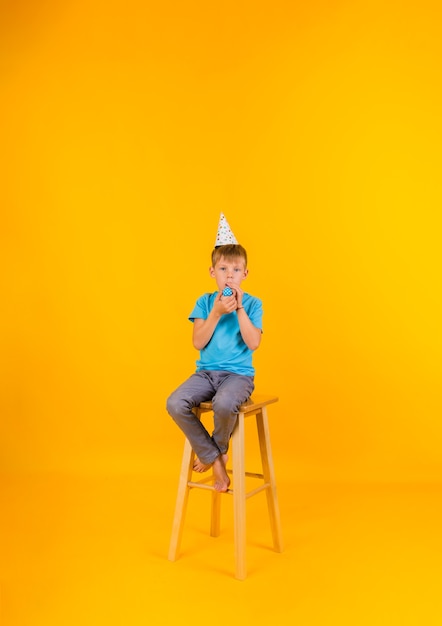 El niño pequeño está sentado en una silla alta con una gorra de vacaciones de papel y con un silbato sobre un fondo amarillo