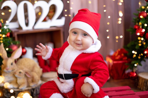 Un niño pequeño está sentado en un trineo bajo los árboles de Navidad. Un gran retrato. Un bebé disfrazado de Papá Noel rojo con un regalo en una caja de regalo. Felices fiestas, año nuevo. Tiempo de Navidad