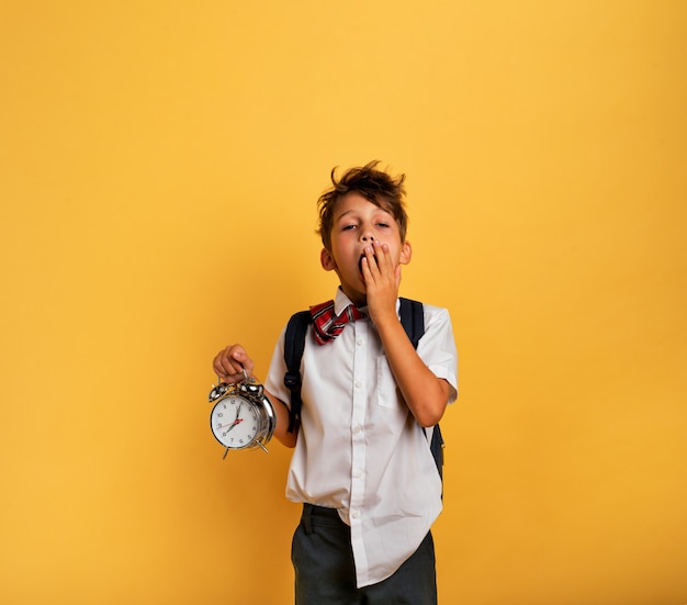 El niño pequeño estudiante con alarma sonando va a ir a la escuela. Fondo amarillo