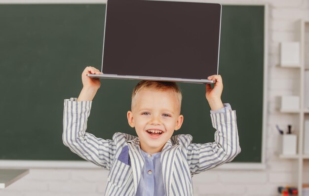 Niño pequeño estudiante sosteniendo la computadora portátil en la cabeza en la clase de la escuela