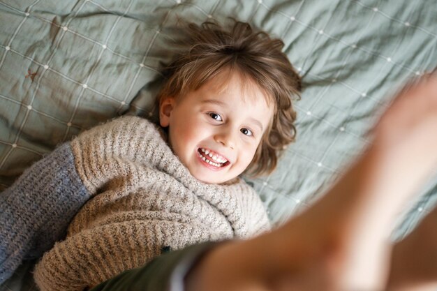 Niño pequeño feliz acostado en el sofá niño extraño acostado en el sofá en casa jugando y saltando piernas lindo niño riendo y mirando a la cámara