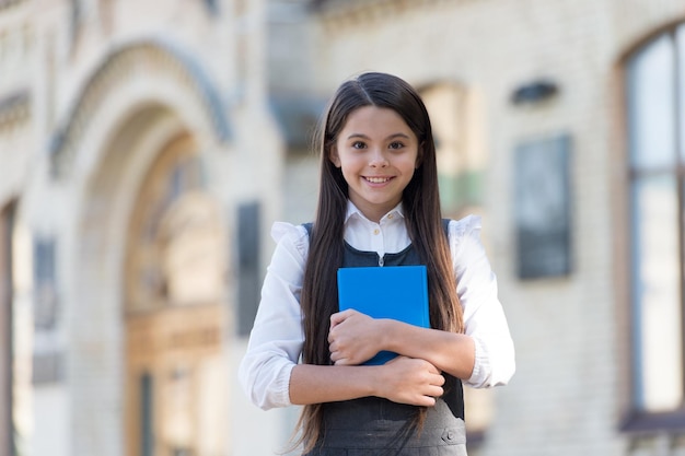 Un niño pequeño feliz con aspecto escolar usa uniforme con un libro de biblioteca al aire libre