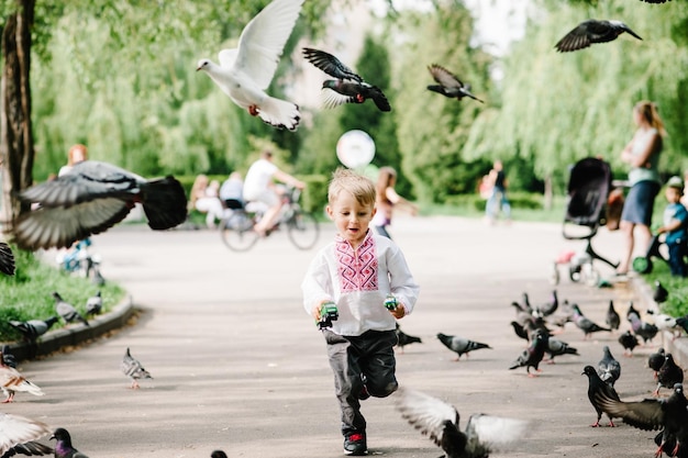 Un niño pequeño y feliz corriendo con una camisa bordada en el fondo, gente de la naturaleza en la calle de la ciudad mirando hacia abajo. El bebé quiere atrapar muchas palomas volando de cerca.