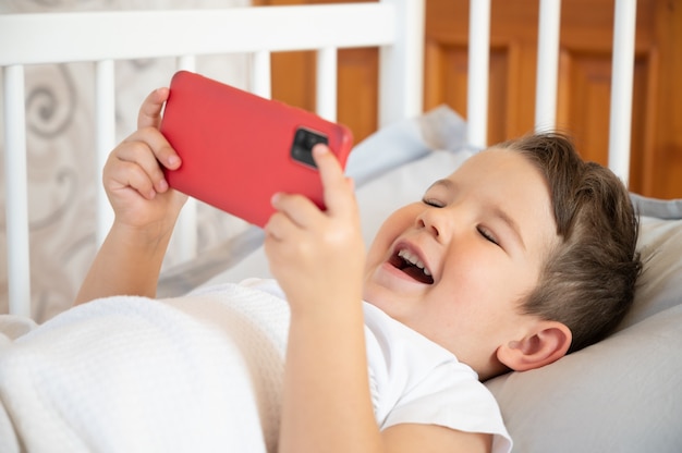 Niño pequeño feliz jugando con un teléfono inteligente en la cama. Concepto de dependencia de Internet. Foto de alta calidad