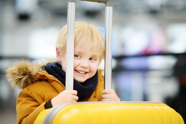 Niño pequeño feliz lindo con la maleta amarilla grande en el aeropuerto internacional antes del vuelo