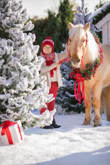 Niño pequeño con gafas acaricia adorable pony con una guirnalda festiva cerca de la pequeña casa de madera y árboles cubiertos de nieve