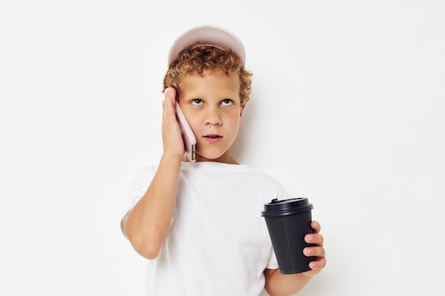 Niño pequeño con una gorra de camiseta blanca con un teléfono en un vaso con un fondo claro de bebida inalterado