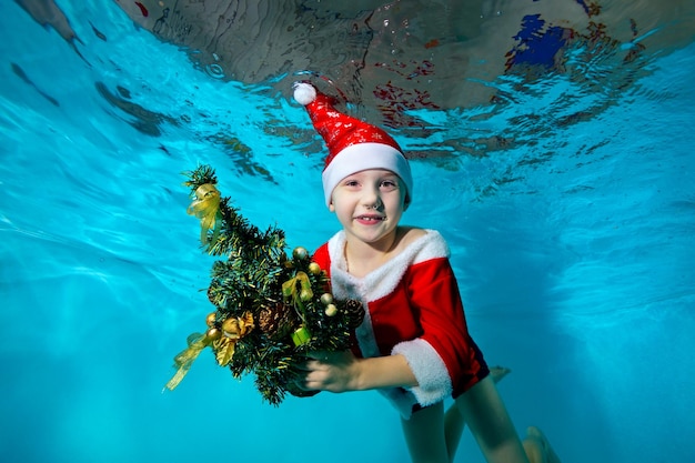 Un niño pequeño con gorra y vestido como Santa Claus nada y posa bajo el agua con un árbol de Navidad