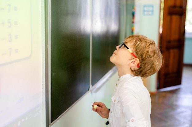 Niño pequeño con grandes gafas negras y camisa blanca de pie cerca de la pizarra de la escuela con un trozo de tiza haciendo cara de pensamiento inteligente