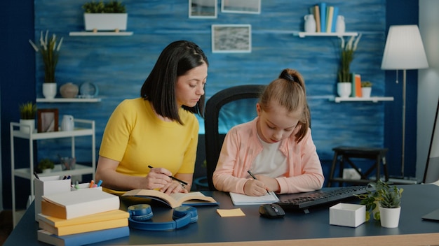 Niño pequeño haciendo la tarea con la madre para la educación remota, usando un libro y un cuaderno en el escritorio. los padres brindan asistencia y ayuda a la niña de la escuela primaria para las tareas de la lección de clase en línea