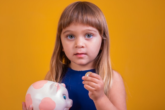 Niño pequeño con hucha sobre fondo amarillo. Niño poniendo monedas en la hucha. Concepto de ahorro y ahorro