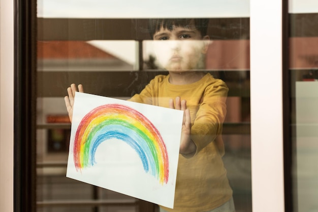 Niño pequeño inspirador que sostiene un dibujo de un arco iris a través de la ventana