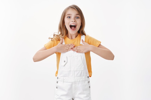 Niño pequeño inteligente feliz emocionado con cabello rubio ojos azules, golpeando con orgullo en el pecho, sonriendo hablando sobre logros personales, fanfarroneando, jurando que puede hacerlo, como voluntario, pararse en la pared blanca