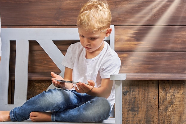 Un niño pequeño con jeans y una camiseta blanca se sienta en un banco de madera con un teléfono inteligente en las manos contra el fondo de una casa de madera Concepto de uso de aparatos por parte de los niños