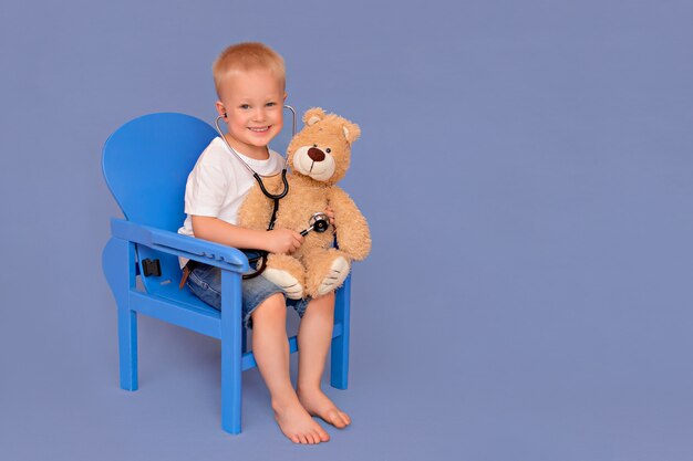 Niño pequeño jugando al doctor y escuchando un oso de peluche con un estetoscopio sobre un fondo azul.