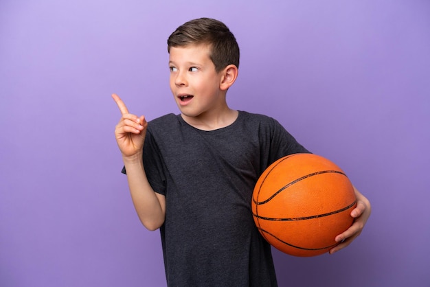 Un niño pequeño jugando baloncesto aislado en un fondo morado con la intención de darse cuenta de la solución mientras levanta un dedo