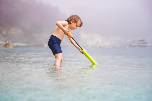 Niño pequeño jugando con un blaster de juguete de agua en el mar en un clima nublado y brumoso Niño pasando las vacaciones de verano en la playa en Europa