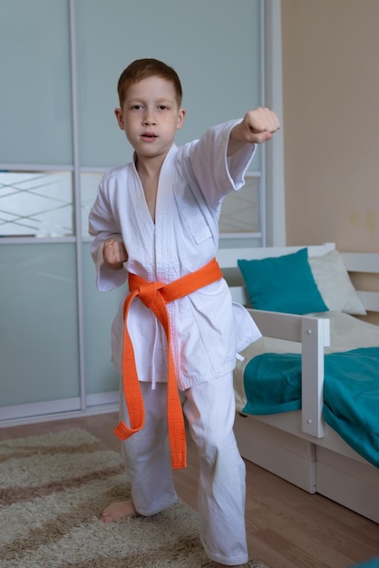 Un niño pequeño en un kimono con un cinturón naranja se para en una posición de combate sobre un fondo claro en casa