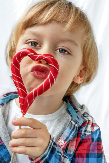 Niño pequeño lindo adentro con las piruletas rojas del caramelo en la forma del corazón, fondo blanco. Hermoso niño come dulces. Día de San Valentín, concepto de amor.