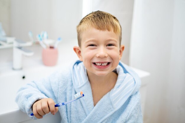 Un niño pequeño y lindo con una bata de baño azul se cepilla los dientes con un cepillo de dientes en casa en un baño brillante salud dental infantil higiene infantil