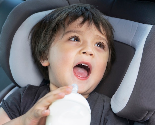 Niño pequeño lindo bebiendo botella de leche en un asiento de automóvil mientras viaja