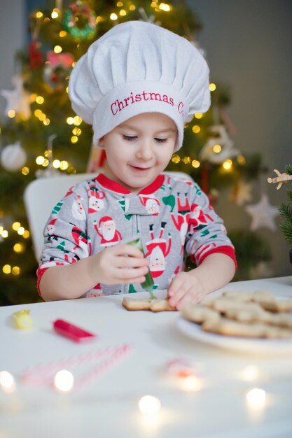 Un niño pequeño y lindo decora las galletas navideñas con un colorido glaseado en una mesa blanca cerca del árbol de Navidad con luces Concepto de cocina navideña