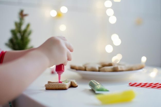 Un niño pequeño y lindo decora las galletas navideñas con un colorido glaseado en una mesa blanca cerca del árbol de Navidad con luces Concepto de cocina navideña