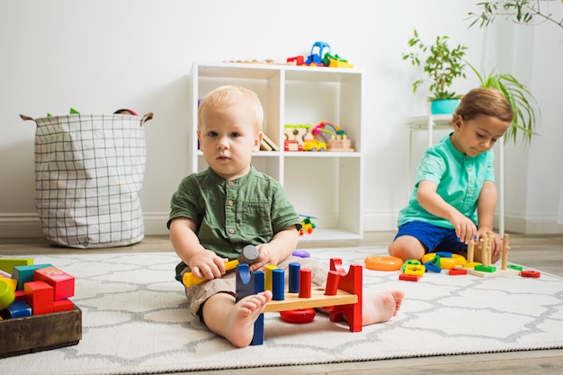 Un niño pequeño lindo está jugando con un martillo de juguete sentado en el piso de su habitación El hermano mayor está jugando con pequeñas pirámides detrás de la acogedora y cómoda habitación de los niños en el fondo