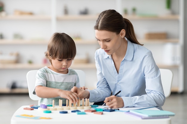 Un niño pequeño y lindo jugando un juego lógico en una psicóloga especializada en gabinetes que toma notas en la oficina