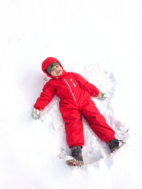 El niño pequeño lindo en un mono rojo de invierno en el invierno en la calle hace que un ángel de la nieve y se divierta.
