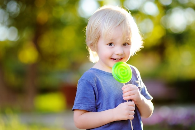 Niño pequeño lindo con la piruleta verde grande. Niño comiendo dulce barra de caramelo. Dulces para niños pequeños. Diversión al aire libre de verano