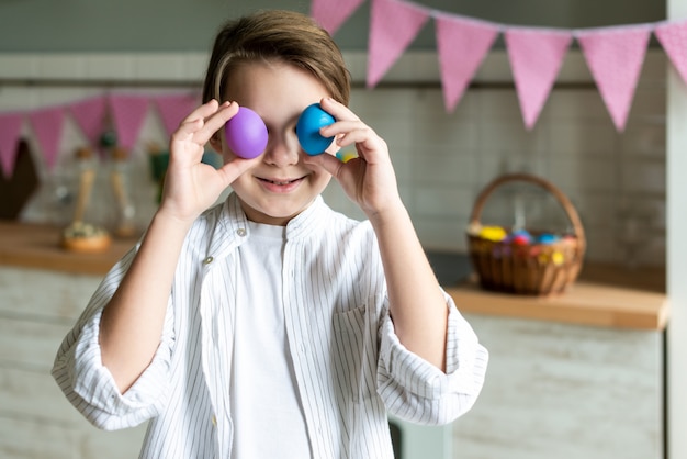 Niño pequeño lindo que sostiene los huevos de Pascua coloreados en vez de ojos. Felices Pascuas .