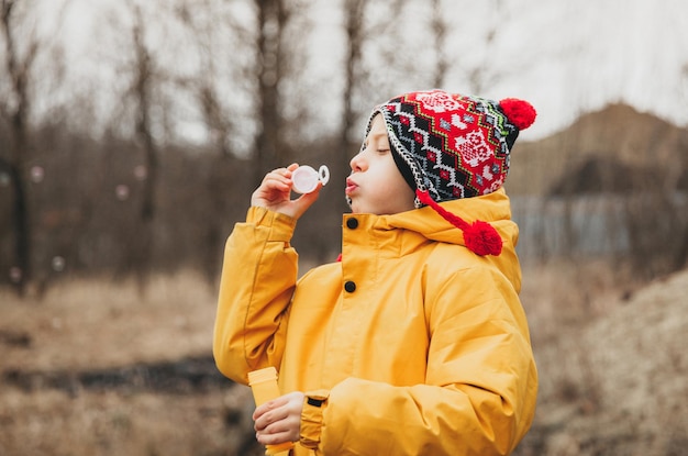 Un niño pequeño y lindo con un sombrero de punto hace pompas de jabón en el parque de otoño.