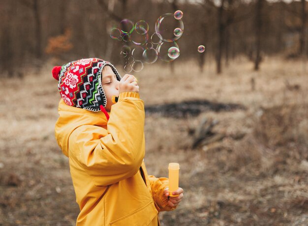 Un niño pequeño y lindo con un sombrero de punto hace pompas de jabón en el parque de otoño.