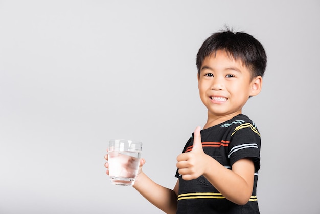 Un niño pequeño y lindo sonríe bebiendo agua fresca de un vaso y muestra el pulgar hacia arriba para una buena señal