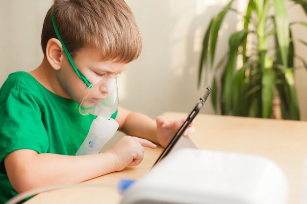 Foto niño pequeño con una máscara para inhalaciones sentado junto al escritorio haciendo inhalaciones con nebulizador en casa