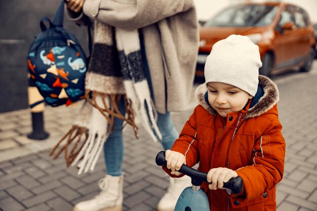 Niño pequeño montando scooter al aire libre e yendo al jardín de infantes mientras su madre lleva