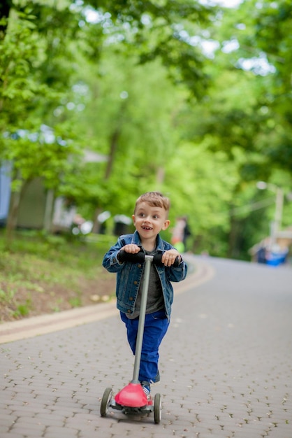 Niño pequeño montando scooter en el parque de la ciudad en un verano Niños haciendo deportes al aire libre Niño feliz jugando con su scooter Niño aprendiendo a montar scooter en un parque Niñez feliz