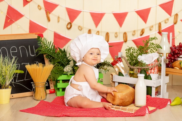 Un niño pequeño una niña de seis meses está sentado en la cocina con la ropa de un cocinero con una hogaza de pan