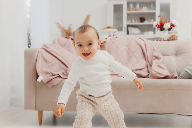 Un niño pequeño, un niño de ojos marrones, está parado cerca del sofá de la habitación. Las emociones, la alegría y la emoción de los niños. Concepto de infancia feliz