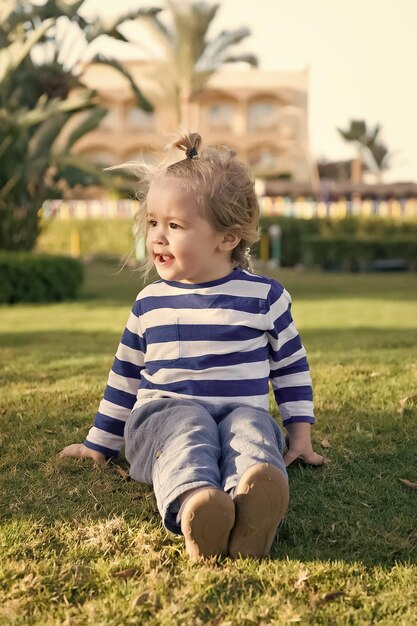 Niño pequeño o niño lindo con cara feliz y cabello rubio en camisa despojada sentado en la hierba verde descalzo al aire libre con sol en el fondo natural