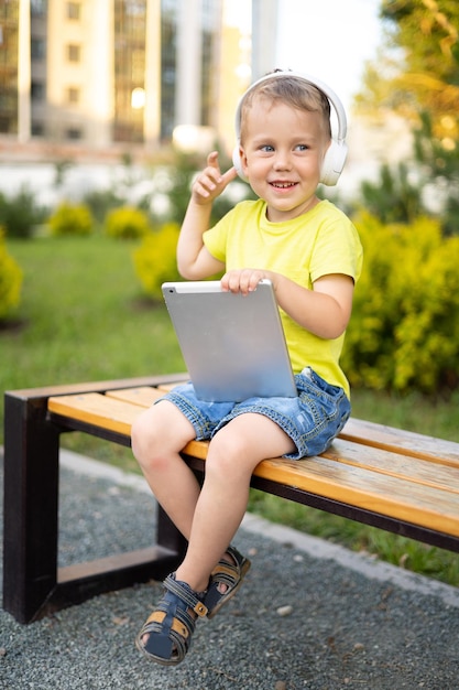 Un niño pequeño con ojos azules y cabello rubio está sentado en un banco del parque con auriculares inalámbricos con una tableta sonriendo riendo jugando