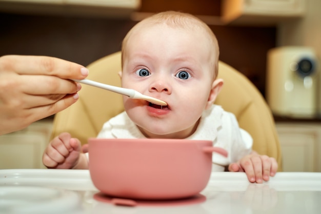 Un niño pequeño con ojos azules es alimentado por una madre con una cuchara los primeros meses de cebo.