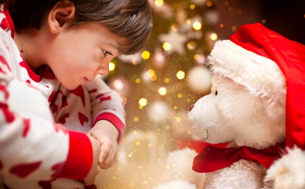 niño pequeño y oso de peluche de cerca en el fondo de las luces de navidad