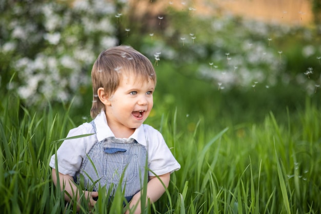 Un niño pequeño con overoles de mezclilla con expresivos ojos azules. Saltando y jugando en la hierba verde alta contra el telón de fondo de un gran arbusto verde y un jardín floreciente.