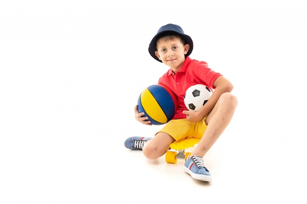 Un niño pequeño en Panamá, una camiseta amarilla, pantalones cortos rojos y zapatillas blancas se sienta en un centavo amarillo y sostiene una pelota de baloncesto multicolor, una pelota de fútbol en blanco y negro y un teléfono en la mano